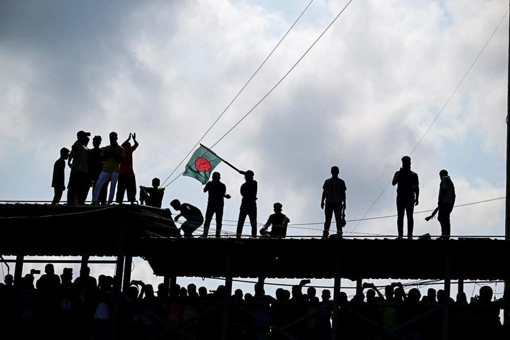 Au Bangladesh, suite aux manifestations de l'été 2024 ayant donné lieu à des crimes de masse, le Tribunal des crimes internationaux reprend du service pour juger les responsables. Mais des rapports s'inquiètent du fonctionnement de la justice locale et de sa politisation. Photo : des manifestants, vus en contre-jour, se tiennent sur une structure en hauteur, dont un brandit un drapeau du Bangladesh.