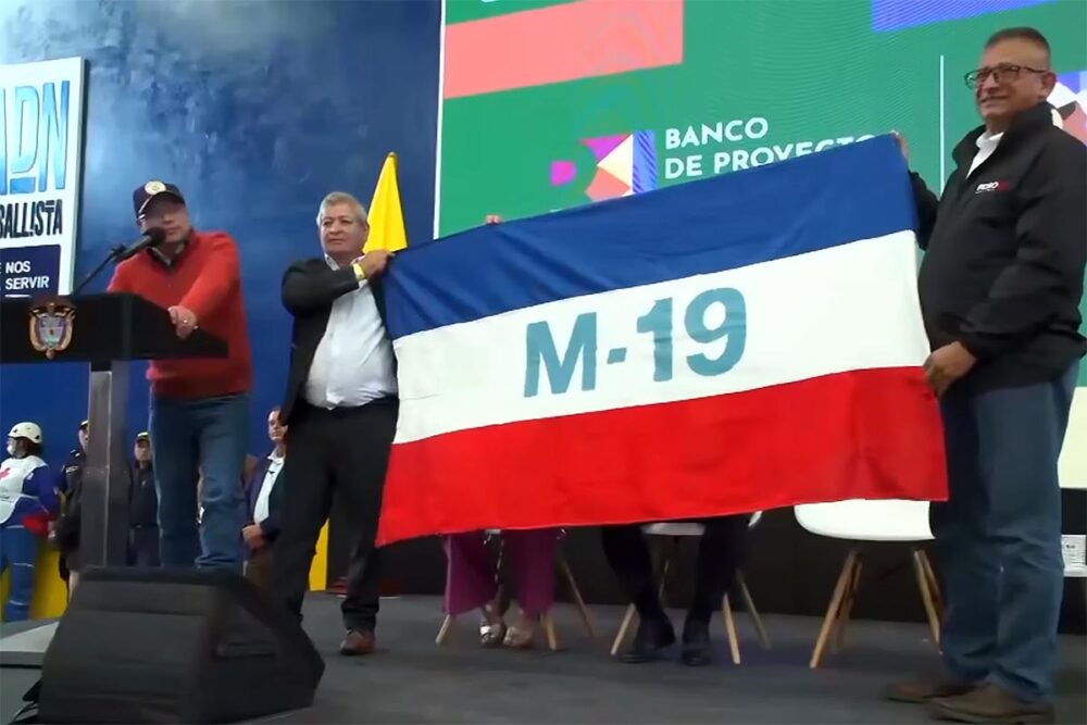 El presidente colombiano Gustavo Petro, entre la guerra y la paz. Foto: Petro pronuncia un discurso en el escenario mientras 2 personas sostienen una bandera del antiguo grupo armado M-19.