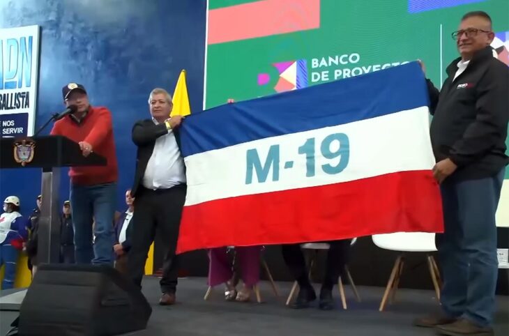Colombian President Gustavo Petro, between war and peace. Photo: Petro delivers a speech on stage while 2 people hold a flag of the former M-19 armed group.