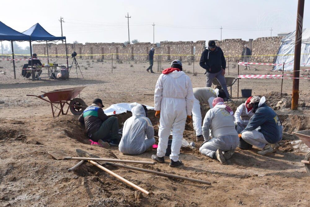 En Irak, une équipe de l'Unitad réalise des fouilles (exhumations) sur les lieux d'une fosse commune. Plusieurs personnes, dont certaines en blouse blanche, s'affairent autour d'un trou creusé sur le sol d'une zone désertique.