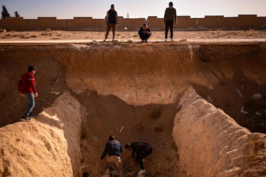 In Syria, after the fall of Assad, the search for the disappeared often begins in mass graves. Photo: People digging at the bottom of a deep mass grave while children watch from above.