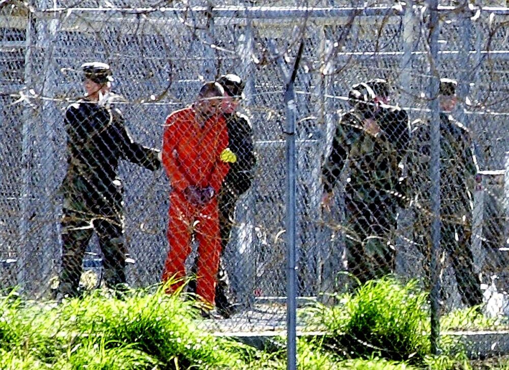 Guantánamo: the American prison (in Cuba) where torture is still practised along with various other violations of human rights (or of the Law in general). Photo: a prisoner in an orange uniform is photographed from a distance, behind barbed wire, surrounded by soldiers.