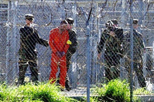 Guantánamo : la prison américaine (à Cuba) où la torture est encore présente tout comme diverses autres violations des droits humains (ou du Droit tout court). Photo : un prisonnier en tenue orange est photographié de loin, derrière des barbelés, et encerclé par des militaires.