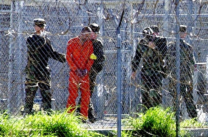 Guantánamo: the American prison (in Cuba) where torture is still practised along with various other violations of human rights (or of the Law in general). Photo: a prisoner in an orange uniform is photographed from a distance, behind barbed wire, surrounded by soldiers.