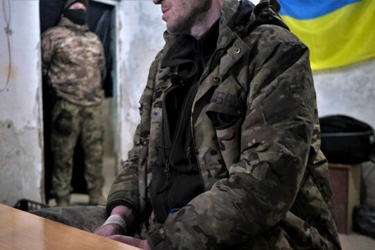 In Ukraine, a military prisoner, sitting on a chair, is guarded by a soldier in the background, in what looks like an interrogation room.