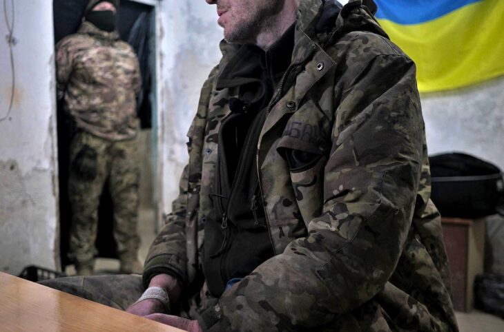 In Ukraine, a military prisoner, sitting on a chair, is guarded by a soldier in the background, in what looks like an interrogation room.